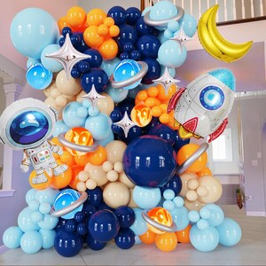 以下が含まれることがあります： 宇宙をテーマにした、鮮やかなバルーンアーチ。青、オレンジ、クリーム色のバルーンに加え、惑星、星、ロケット、宇宙飛行士、三日月型のホイルバルーンが配置されています。宇宙をテーマにしたパーティーに最適です。