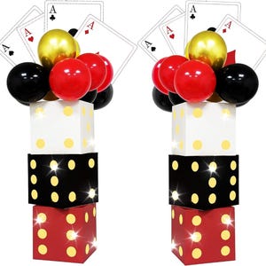Puede incluir: Dos torres decorativas con cajas en forma de dados apiladas en blanco, negro y rojo, rematadas con racimos de globos dorados, rojos y negros y grandes cartas de juego que muestran los ases de picas, corazones y diamantes.