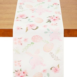 Puede incluir: Camino de mesa blanco de Pascua con un patrón repetido de conejitos rosas, huevos de Pascua y flores rosas. El camino de mesa es rectangular y está hecho de una tela clara, perfecto para la decoración primaveral. El camino de mesa está colocado sobre una mesa de madera.