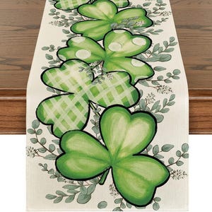 Puede incluir: Camino de mesa del Día de San Patricio con grandes tréboles verdes con lunares blancos y patrones de cuadros. Los tréboles están rodeados de hojas verdes y pequeñas flores blancas sobre un fondo crema.
