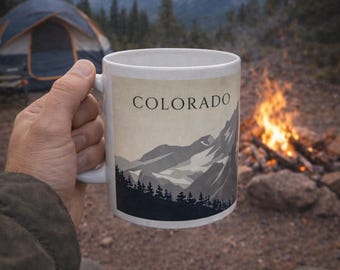 May include: White ceramic mug with a Colorado mountain landscape design. The mug features a grayscale illustration of mountains and trees, with the word "COLORADO" at the top. The mug is held outdoors near a campfire and tent.