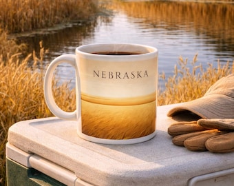 May include: A white coffee mug with the word "NEBRASKA" printed on it, featuring a landscape scene of golden fields. The mug sits on a cooler next to a hat and gloves, with a river and trees in the background.