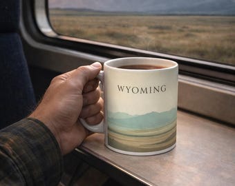 May include: A white ceramic mug filled with a dark beverage, featuring a landscape design and the word "WYOMING." The mug is held near a train window, with a scenic view of mountains and a sunset in the background.