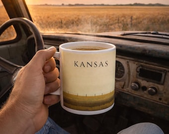 May include: A white ceramic mug with the word "KANSAS" printed on it, held in a person's hand. The mug features a landscape design of a field and a fence. The background shows a sunrise over a field.