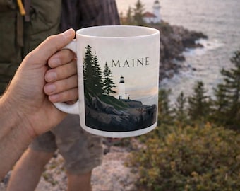 May include: White ceramic mug featuring a coastal Maine scene with a lighthouse, trees, and the word "MAINE." The mug is held by a person hiking outdoors, with a backpack and scenic background of the ocean and rocky coastline.
