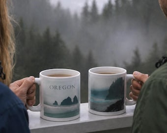 May include: Two white ceramic mugs with landscape designs and the word "OREGON" printed on one. The mugs are filled with a dark liquid, likely coffee or tea. The background shows a misty coastal scene.