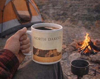 May include: A white ceramic mug with the words "NORTH DAKOTA" and a landscape image, filled with dark coffee. The mug is held outdoors near a campfire and a tent, suggesting a camping theme.