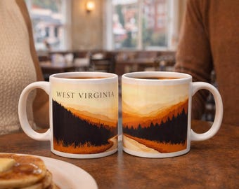 May include: Two white ceramic mugs with the text "WEST VIRGINIA" and a landscape design. The design features a mountain range in shades of orange, yellow, and brown, with a dark forest at the base. The mugs are filled with a dark liquid.