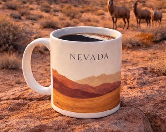 May include: A white ceramic mug filled with coffee, featuring a landscape design with the word "NEVADA". The mug is set on a reddish-brown rock, with a desert scene and bighorn sheep in the background.