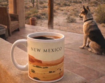 May include: A white ceramic mug filled with dark coffee, featuring the words "NEW MEXICO" and a desert landscape design. A dog sits in the background. The mug is on a stone surface.