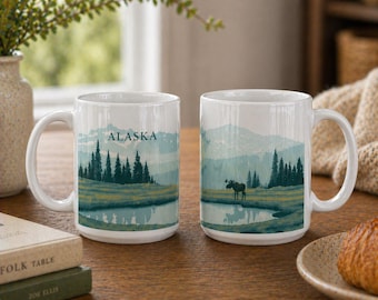 May include: Two white ceramic mugs with a landscape design. The design features a lake, evergreen trees, and mountains in shades of blue and green. The word "ALASKA" is printed on the mugs.