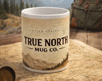 May include: A white ceramic mug with the text "TRUE NORTH MUG CO." and a mountain illustration. The mug is on a vintage map with a compass, and a backpack is in the background. The mug is designed for outdoor enthusiasts.