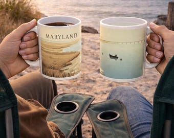 May include: Two white ceramic mugs with landscape designs are held by people sitting in camping chairs. One mug says "Maryland" and shows a coastal scene. The other mug has a boat on the water. The mugs are filled with coffee.