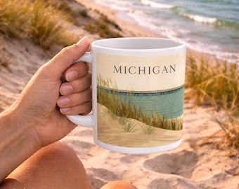 May include: White ceramic mug with a beach scene and the word "MICHIGAN" printed on it. The mug features a sandy beach, water, and tall grass. The mug is held by a person on a sandy beach with the ocean in the background.