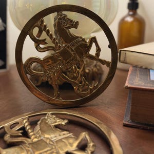 Könnte beinhalten: Zwei Untersetzer aus Messing mit Karussellpferden. Jeder Untersetzer zeigt ein detailliertes Karussellpferd-Design in einem kreisförmigen Rahmen. Das Messing hat einen warmen, goldenen Farbton. Die Untersetzer werden auf einer Holzoberfläche präsentiert.