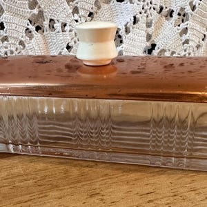 May include: A rectangular glass butter dish featuring a clear, vertically ribbed base and a polished copper lid. The lid is topped with a small, solid white ceramic knob handle, providing a contrast to the metallic finish of the cover.