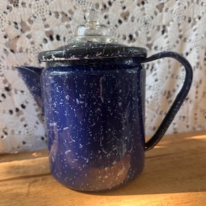 May include: A dark blue enamel coffee pot with a white speckled pattern, featuring a curved handle, a side spout, and a clear glass lid with a pointed finial. The cylindrical body has a smooth finish and a dark rim around the top edge.