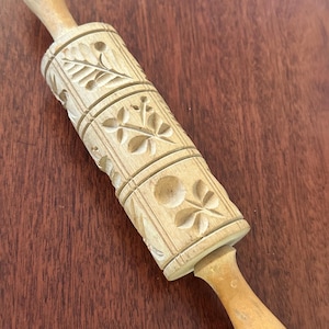 May include: A light-colored wooden rolling pin featuring a cylindrical barrel with deeply carved botanical patterns, including leaves, berries, and floral motifs. The tool has two smooth, tapered handles on either end for manual rotation.