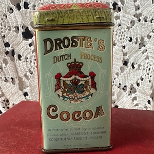 May include: A rectangular metal tin for Droste's Dutch Process Cocoa with a light green front panel featuring a red and gold crest, black text, and a red lid. The text reads: Droste's Dutch Process Cocoa, is manufactured by a special process which INCREASES THE MINERAL CONSTITUENTS ABOUT 3 PERCENT, MADE IN HOLLAND.