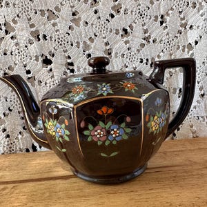 May include: A dark brown ceramic teapot with a hexagonal body, a matching lid, and a curved handle. The teapot features hand-painted floral motifs in orange, blue, yellow, and green, accented with thin gold lines along the edges of the panels.