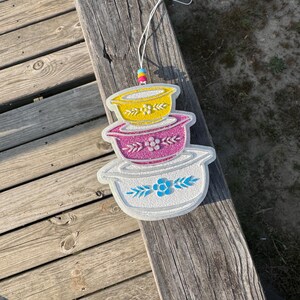May include: A decorative hanging ornament featuring a stack of three colorful bowls. The top bowl is yellow, the middle is pink, and the bottom is white. Each bowl has a floral design. The ornament hangs from a white string.