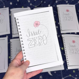 May include: Several white spiral-bound notebooks with the text "Issue ZERO" in a handwritten style. A pink flower illustration is above the text. The notebooks are on a blue patterned surface.