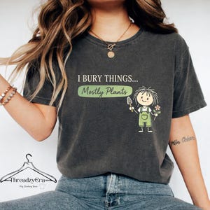 Könnte beinhalten: Dunkelgraues T-Shirt mit dem Text "I BURY THINGS... Mostly Plants" in Weiß und Grün. Ein Cartoon-Gärtner mit einer Kelle und einer Blume ist ebenfalls abgebildet. Das Shirt ist ein lässiger, bequemer Stil.