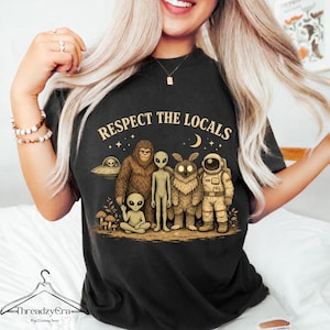 Puede incluir: Camiseta negra con la frase "Respect the Locals" sobre un gráfico de un OVNI, Bigfoot, extraterrestres, un Mothman y un astronauta. El diseño utiliza tonos marrones, beige y blancos.