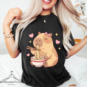 Peut inclure: T-shirt noir avec un capybara de dessin animé mangeant des ramen avec des baguettes. Le capybara porte un nœud rose et est entouré de cœurs roses et d'étoiles jaunes. Le t-shirt porte le nom de la marque "ThreadzyEra" en bas.