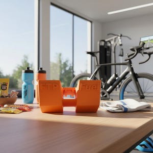 Op de afbeelding: Een oranje FTP Fuel-houder met de tekst "FTP Fuel" staat op een houten tafel, samen met waterflessen, energiegels en een kom met snacks. Een hometrainer en fitnessapparatuur zijn op de achtergrond te zien.