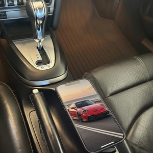 May include: A smartphone resting on the black leather center console of a vehicle interior. The phone screen displays a high-resolution image of a red sports car driving on a coastal road. The car interior features a silver gear shifter.