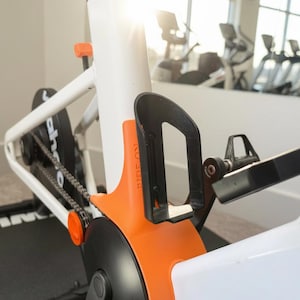 May include: A bright orange Zwift Ride Crank Protector mounted on a white stationary bike frame. The protector features a black bottle cage attached to its side and the text Ride On embossed vertically on the orange surface near the frame.