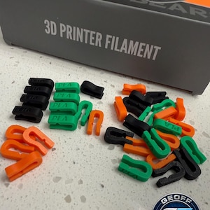 May include: A collection of black, green, and orange 3D printer filament clips scattered on a white surface. The clips are in various shapes and sizes. A box labeled "3D PRINTER FILAMENT" is in the background.