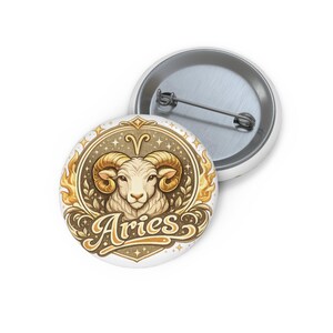 Pode incluir: Botão branco com o signo do zodíaco de Áries. O design apresenta uma cabeça de carneiro com chifres dourados, rodeada por uma borda decorativa e a palavra "Aries" em uma fonte estilizada.