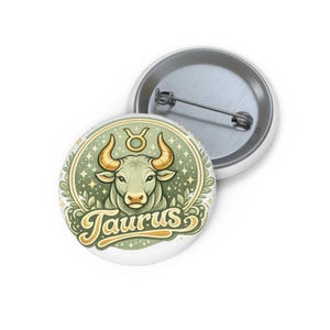 Puede incluir: Un botón blanco con una ilustración verde y dorada de la cabeza de un toro, el símbolo del zodiaco Tauro y la palabra "Taurus". Un segundo botón es visible en el fondo.