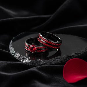 Puede incluir: Dos anillos negros con piedras rojas. Un anillo presenta una gran piedra roja en forma de corazón, mientras que el otro tiene múltiples filas de piedras rojas más pequeñas. Los anillos se exhiben sobre una superficie oscura y reflectante sobre un fondo negro.