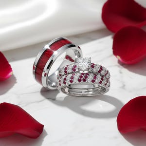 Peut inclure: Deux alliances et un ensemble de bagues assorties sont présentés. La première bague a une incrustation en bois rouge et un anneau argenté. Le deuxième ensemble de bagues a un diamant en forme de cœur et des accents de rubis. Des pétales de rose rouges sont éparpillés.