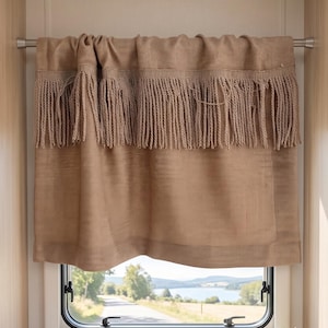 May include: A tan window curtain with a fringed valance. The curtain is made of a woven fabric and hangs above a window with a view of a road and lake. A small potted plant and a mug sit on the windowsill.