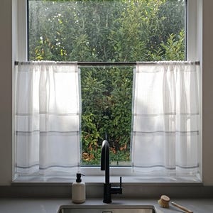Könnte beinhalten: Weiße Küchenvorhänge mit grauen horizontalen Streifen hängen vor einem Fenster. Eine schwarze Armatur und ein Edelstahlbecken befinden sich im Vordergrund. Ein Seifenspender und eine Spülbürste stehen auf der Arbeitsplatte.