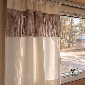 Op de afbeelding: Crèmekleurige gordijnen met een decoratieve franje. De gordijnen hangen voor een raam, met uitzicht op bomen en een recreatievoertuig op de achtergrond. De gordijnen zijn gemaakt van een lichtgekleurde stof.
