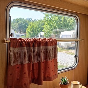 May include: A window in a camper with a rust-colored curtain with fringe. A gold-colored rod holds the curtain. A small potted succulent and a mug sit on a wooden table. The window looks out onto a sunny day.