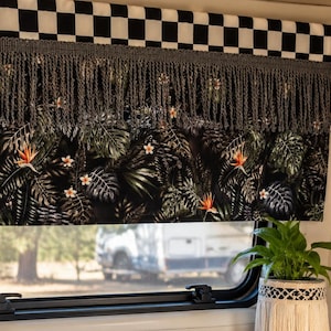Puede incluir: Una cenefa decorativa con una parte superior a cuadros en blanco y negro, flecos grises y un estampado floral tropical. La cenefa está instalada sobre una ventana, con una planta en una maceta de macramé visible.