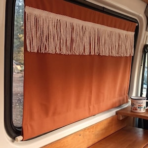 May include: A burnt orange window curtain with a cream fringe, inside a van. The curtain is next to a window with a view of trees and a campsite. A small wooden shelf holds a mug with the word "Travel".
