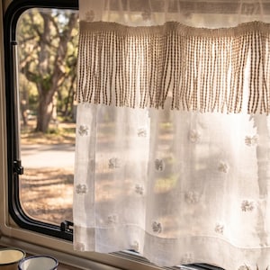 Könnte beinhalten: Weißer Leinen-Vorhang mit Fransenbesatz und floraler Stickerei. Der Vorhang hängt vor einem Fenster mit Blick auf Bäume und eine Straße. Der Vorhang befindet sich in einem Wohnmobil, mit Tassen und einer kleinen Pflanze.