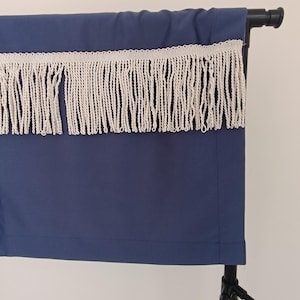 May include: A navy blue fabric panel with a cream-coloured fringe trim, displayed on a black stand. The fringe hangs below the top edge of the panel. The fabric is smooth and appears to be a solid colour.