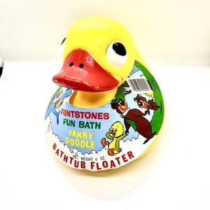 Peut inclure: Un flotteur de bain vintage jaune en forme de canard, avec un bec rouge et des yeux noirs. Le produit porte une étiquette avec le texte "Flintstones Fun Bath Yakky Doodle Bathtub Floater" et des personnages de dessins animés. Poids net 170 grammes.
