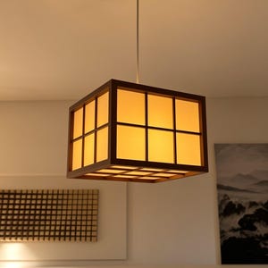 May include: A square pendant light fitting featuring a dark brown wooden frame with a grid pattern and translucent, warm yellow panels. The lamp is suspended from the ceiling by a single thin rod and emits a soft, diffused glow from within.