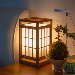 Könnte beinhalten: Eine quadratische Tischlampe aus Holz mit Gitterdesign, die ein warmes Leuchten ausstrahlt. Die Lampe steht auf einer Holzoberfläche, neben einer kleinen Topfpflanze und einem Kaktus. Das Design der Lampe erinnert an japanische Shoji-Bildschirme.