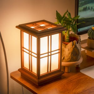 Könnte beinhalten: Quadratische Holzlaternenlampe mit dunkelbraunem Rahmen und Gittermuster oben. Die Lampe hat vertikale und horizontale Stäbe an den Seiten, durch die das Licht durch weiße Paneele scheint. Sie steht auf einer Holzoberfläche. Die Lampe misst etwa 20 cm.