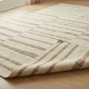 Op de afbeelding: Beige en bruin gestreept vloerkleed met een opgerolde rand, met een natuurlijke vezeltextuur. Het vloerkleed heeft meerdere bruine strepen over het oppervlak, waardoor een eenvoudig maar elegant ontwerp ontstaat. Het vloerkleed ligt op een lichtgekleurde houten vloer.